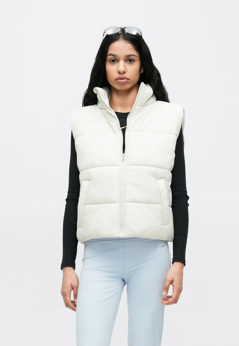 Gepolsterte, ärmellose Weste in hellem Creme mit hohem Kragen, Reißverschluss vorne und Seitentaschen, getragen über einem schwarzen Langarmshirt und blauen Leggings.