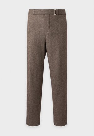 Jeune homme portant un pull en maille marron sur une chemise foncée, un pantalon marron avec une ceinture et des mocassins noirs en cuir à pampilles, debout devant un fond blanc.
