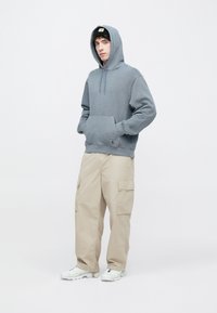 Szara bawełniana bluza z kapturem, z przednią kieszenią i sznurkiem do regulacji kaptura, zestawiona z beżowymi spodniami typu cargo z bocznymi kieszeniami oraz białymi sneakersami.