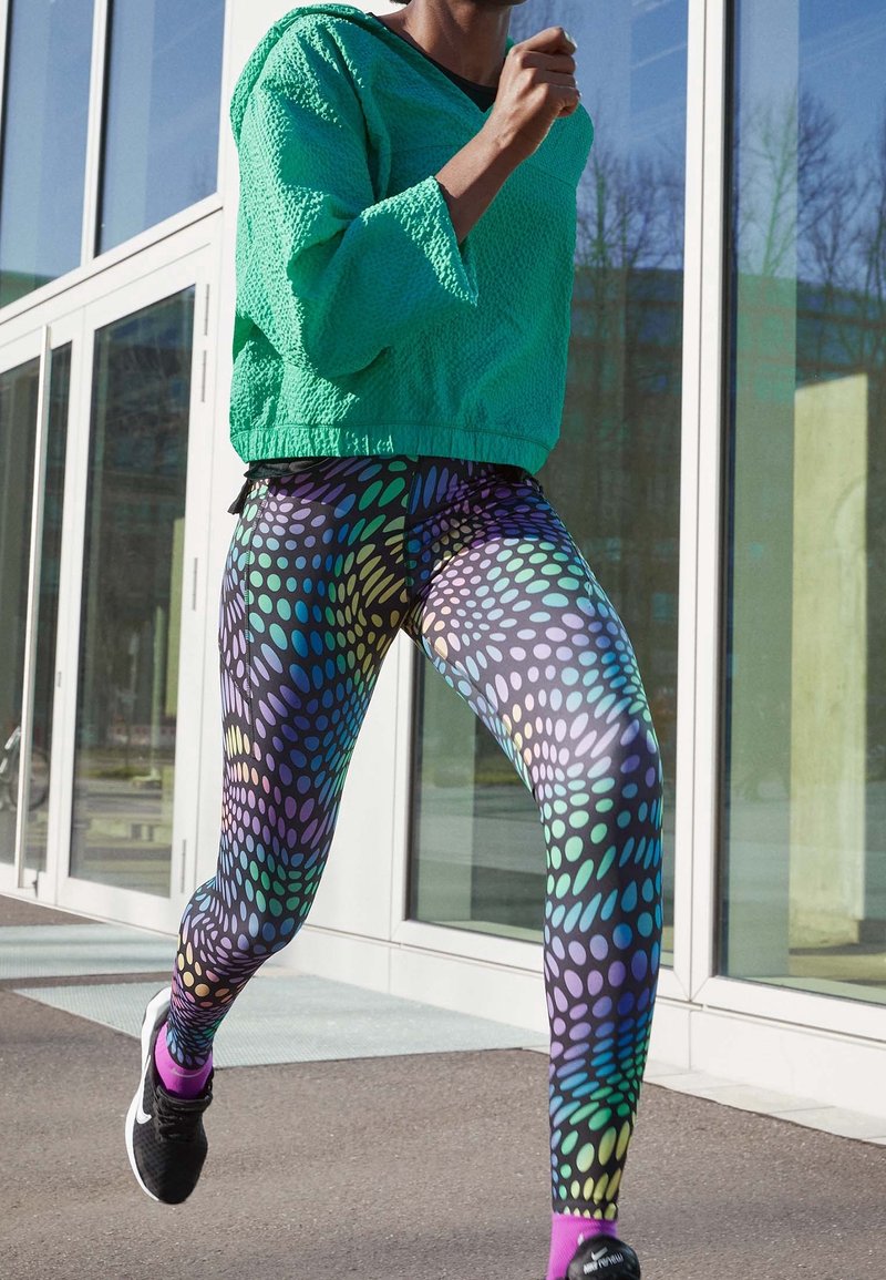 Giacca a vento verde con texture abbinata a leggings colorati e stampati con pois in tonalità di blu, verde e viola. Sneakers nere.