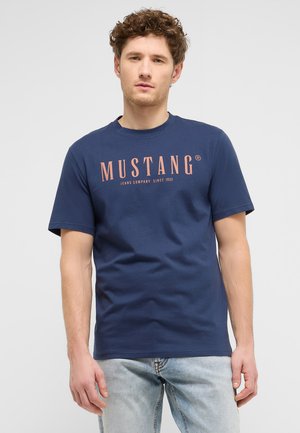 Junger Mann mit lockigem Haar, der ein dunkelblaues Mustang-T-Shirt und hellblaue Jeans trägt, steht vor einem schlichten grauen Hintergrund.