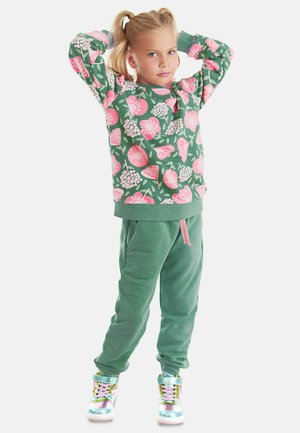 Grüner Sweatshirt mit floralem Muster und pinken Akzenten, kombiniert mit einfarbigen grünen Jogginghosen. Model trägt schimmernde, mehrfarbige High-Top-Sneaker.