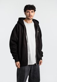 Sort zip-up hoodie lavet af blødt stof med en rummelig pasform, der har en stor hætte, forlommer og ribbede manchetter, lagdelt over en hvid t-shirt.