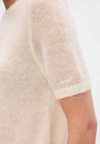 Pletený sveter s krátkym rukávom v jemnej svetloružovej farbe s priesvitnou textúrou. Obsahuje rebrovaný biely manžet a malý detail loga.
