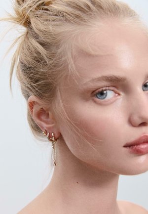 Goldene Ohrringe mit verschiedenen Designs, darunter Creolen und ein fallender Kettenanhänger, getragen von einer hellhäutigen Person mit blonden Haaren.