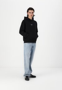 Svart hettegenser med frontlomme og logo, kombinert med lyseblå baggy jeans og svarte sneakers med hvite striper. Enkel hvit bakgrunn.