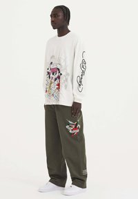 Wit longsleeve shirt met kleurrijke graphic van een schedel en saxofoon; groene broek met geborduurd rood en groen ontwerp; witte sneakers.
