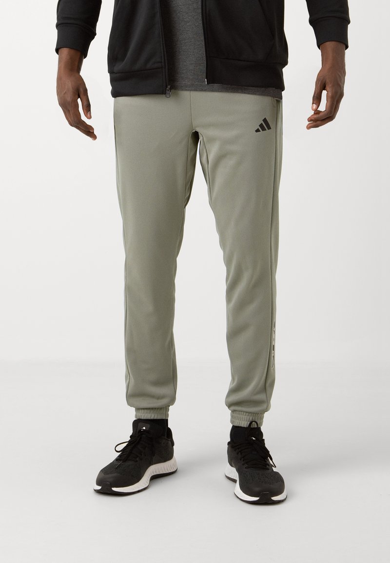Szare-zielone legginsy sportowe z elastycznymi mankietami, bocznymi paskami i logo Adidas. W zestawie z czarnymi butami biegowymi z białymi podeszwami.