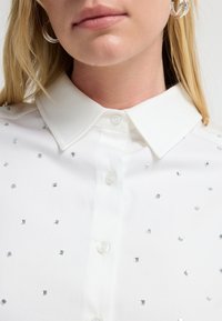 Blusa blanca de botones con cuello clásico, que presenta lentejuelas plateadas cuadradas dispersas y una textura suave. Primer plano del escote.
