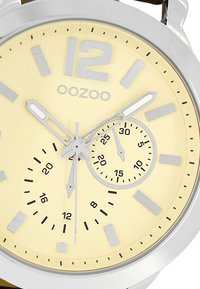 Montre argentée avec un boîtier rond, un cadran jaune texturé, des index horaires gris et deux sous-cadrans pour les secondes et les minutes. Nom de marque "OOZOO" affiché.