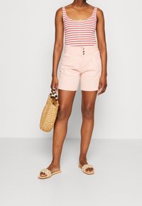 Body rouge et blanc rayé avec des boutons, short rose clair cintré avec des plis à l'avant, sac en osier beige et sandales de couleur claire.