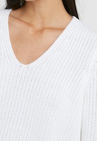 Biały sweter z dzianiny z dekoltem w serek, o luźnym kroju, z teksturowanym wzorem i prążkowanymi detalami na dekolcie oraz rękawach.