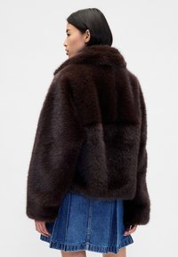 Giacca marrone in pelliccia sintetica con colletto ampio, spalle arrotondate e vestibilità squadrata, abbinata a una gonna di jeans. La texture è morbida e densa.