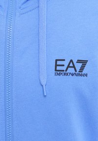 Svetlo modrá zateplená mikina na zips vyrobená z mäkkého materiálu s hladkou textúrou. Obsahuje čierne logo EA7 Emporio Armani na ľavej strane hrudníka a pletené šnúrky.