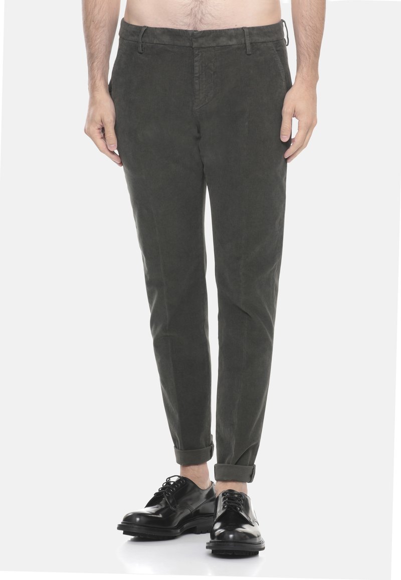 Uomo che indossa pantaloni in velluto a coste verde scuro arrotolati alla caviglia e scarpe eleganti nere lucidate, in piedi su uno sfondo bianco.