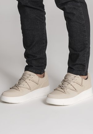 Persoon die beige lage sneakers met witte zolen en zwarte spijkerbroek draagt, staand op een lichtgrijze ondergrond.