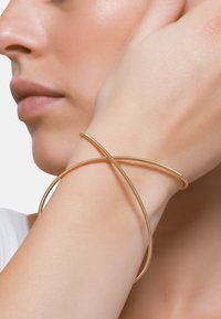 Bangle armbånd i gullfarget metall, med en stilren, minimalistisk design og to sammenvevde, jevne bånd. Overflaten er blank.
