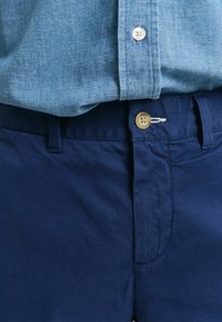 Marineblauwe shorts van gladde stof, voorzien van een knoopsluiting met een beige knop en een voorkeurszak. Blauw overhemd met knopen eroverheen.