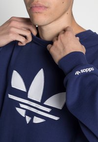 Mörkblå sweatshirt med en stor vit Adidas trefoil-logotyp och tre horisontella ränder, med ribbad mudd och halsringning.