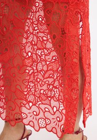 Roter Spitzenrock mit floralen Mustern und einem offenen Design. Verfügt über einen seitlichen Schlitz und einen gewellten Saum, der die filigranen Details betont.