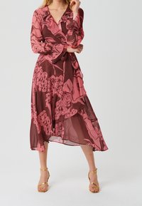 Mujer con un vestido envolvente de flores en tonos rosa y marrón, de manga larga, y sandalias de tacón alto doradas con tiras, posando contra un fondo liso.