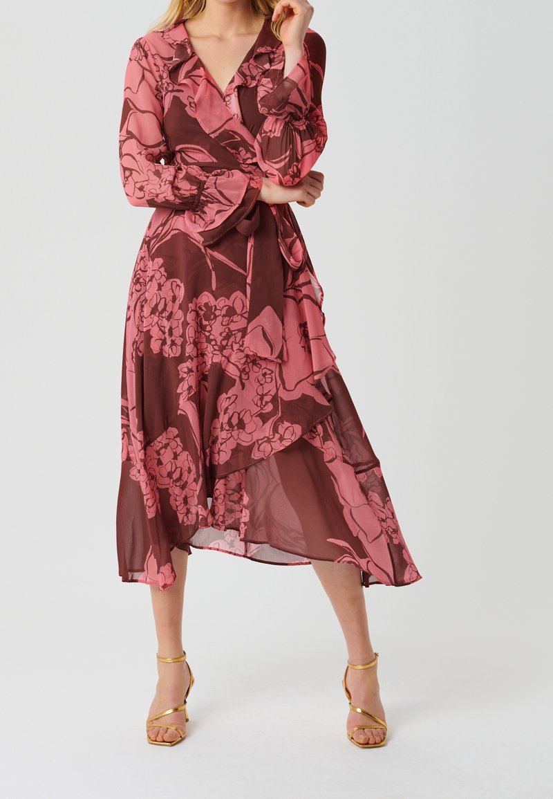 Mujer con un vestido envolvente de flores en tonos rosa y marrón, de manga larga, y sandalias de tacón alto doradas con tiras, posando contra un fondo liso.