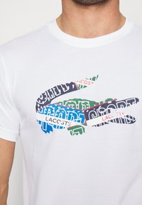 White cotton t-shirt featuring a multicolored graphic of a crocodile logo with text elements in blue, green, red, and black.