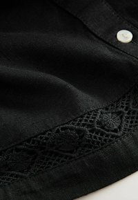 Top in tessuto nero con una superficie liscia, caratterizzato da un dettaglio in crochet decorativo lungo l'orlo inferiore e una chiusura con un singolo bottone.