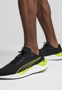 Zapatillas deportivas negras con una parte superior de tela texturizada, que presentan acentos amarillos vibrantes y una entresuela blanca. Diseño con cordones y con un cuello acolchado.