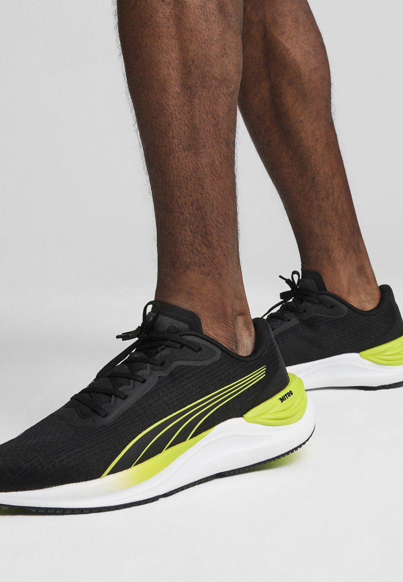 Zapatillas deportivas negras con una parte superior de tela texturizada, que presentan acentos amarillos vibrantes y una entresuela blanca. Diseño con cordones y con un cuello acolchado.