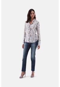 Blumenhemd mit Knöpfen aus leichtem Stoff mit weißem Grund, das lila und blaue Muster aufweist. Getragen mit dunkelblauen Jeans und hellblauen Pumps.