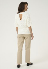 Pull en maille blanche à manches courtes avec un découpage dans le dos, associé à un pantalon beige et des ballerines noires, sur un fond clair.