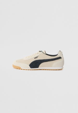 Baskets Puma Arizona beige avec bande latérale noire, semelle en gomme et design à lacets, vues de côté extérieur sur fond blanc.