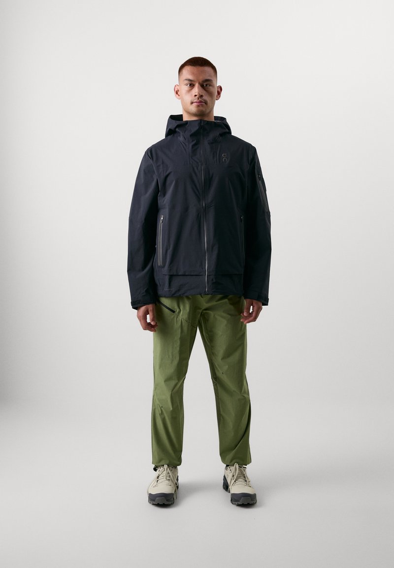 Chaqueta impermeable azul oscuro con capucha, frente con cremallera y bolsillos, combinada con pantalones verde claro y zapatos de senderismo beige.