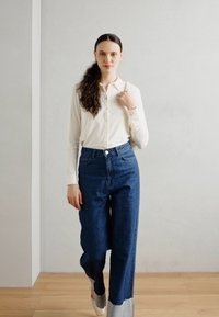 Vit skjorta med lång ärm, kombinerad med högmidjade, vida blå denimjeans med en kontrasterande fåll. Platta skor.