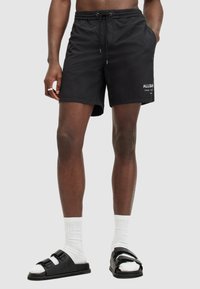 Black swim shorts with an elastic waistband, drawstring closure, and side pockets. Features white logo text on the bottom hem.