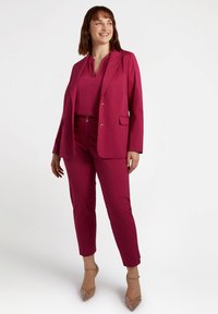 Completo fucsia con giacca sartoriale e pantaloni affusolati. Tessuto liscio, bottoni dorati e una blusa abbinata. Silhouette chiara e moderna.