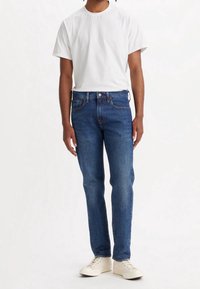 Vaqueros de denim azul con corte recto, con cierre de botones y cinco bolsillos, combinados con una camiseta blanca lisa de cuello redondo y zapatillas beige.