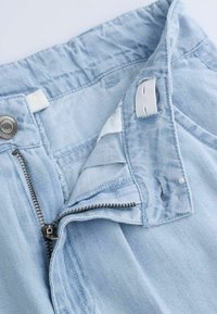 Jasnoniebieskie dżinsy pokazujące rozpiętą suwak, zatrzask i szczegóły wewnętrznej tkaniny. Charakteryzują się gładką fakturą i wyraźną konstrukcją pasa.