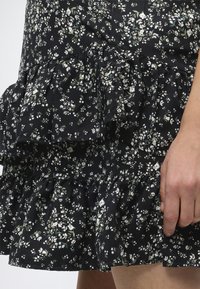 Μαύρη φούστα με στρώσεις και μικρό λευκό ανθισμένο σχέδιο, σκουληκότρυπες ραφές, και κοντινή εικόνα ενός χεριού που ξεκουράζεται στο πλάι.