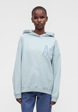 Jeune personne aux cheveux courts portant un sweat à capuche bleu clair avec de grandes lettres "AB" sur la poitrine et un jean bleu.