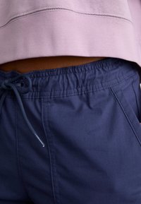 Shorts bleu marine à cordon de serrage en tissu doux, avec une taille élastique et des détails de surpiqûres minimalistes. Partiellement couverts par un haut rose.