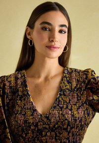 Gemustertes Kleid mit tiefem V-Ausschnitt, das reichhaltige blaue, gelbe und lila Texturen aufweist. Das Model trägt goldene Creolen.