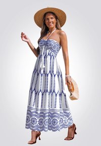 Mujer en un vestido maxi blanco sin mangas con patrones azules, llevando un sombrero de paja de ala ancha, tacones marrones y sosteniendo un bolso de mimbre.