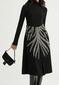 Schwarzes Kleid mit langen Ärmeln, das vorne ein weißes Blattmuster und zwei Seitentaschen aufweist. Dazu eine kleine, strukturierte schwarze Tasche.