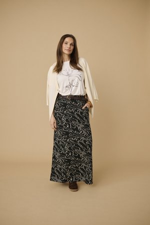 Kvinde iført hvid cardigan over hvid top med blomsterprint, sort mønstret maxiskørt og brune sko, står med hånden i lommen.