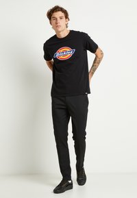 T-shirt di cotone nera con collo rotondo e un colorato logo "Dickies" stampato al centro. Pantaloni neri slim-fit e sneakers nere con accenti gialli.