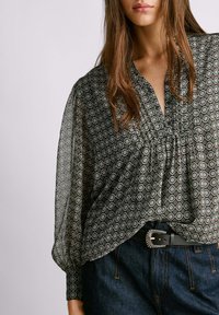 Blusa nera a fantasia con scollatura a V, giromanica plissettato e maniche lunghe a sbuffo. Abbinata a jeans in denim blu a vita alta e una cintura nera.