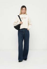 Giovane donna che indossa un sweater floreale color crema, jeans larghi blu scuro, scarpe nere e porta una borsa a spalla nera su uno sfondo bianco.