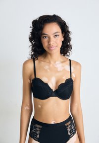 Melna mežģīņu push-up krūšturis ar apmalēm, ar polsterētām krūšu kausiem un regulējamām siksnām, kopā ar augstiem melniem apakšbiksēm ar mežģīņu akcentiem.